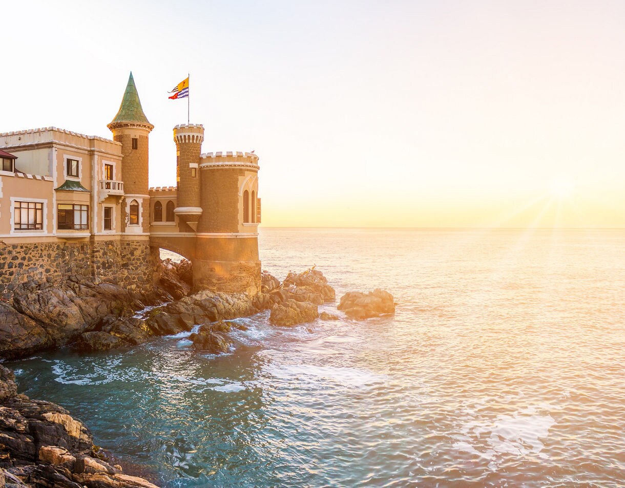 Sunset light washes over Castillo Wulff, a turreted seaside mansion perched on coastal rocks above gentle Pacific waves.