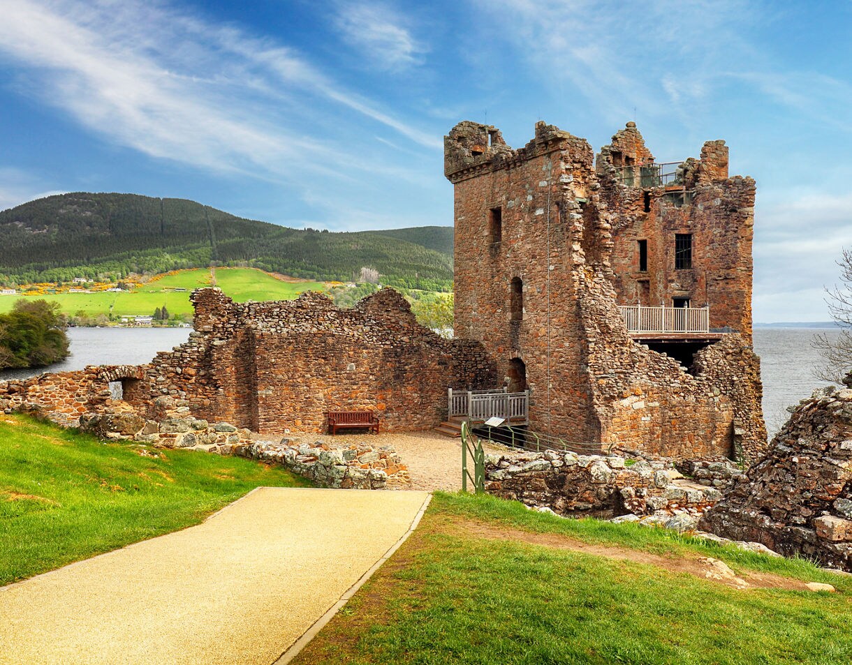 Stone ruins of Urquhart Castle with crumbling walls and a tall tower, set beside Loch Ness with green hills and forests in the distance.