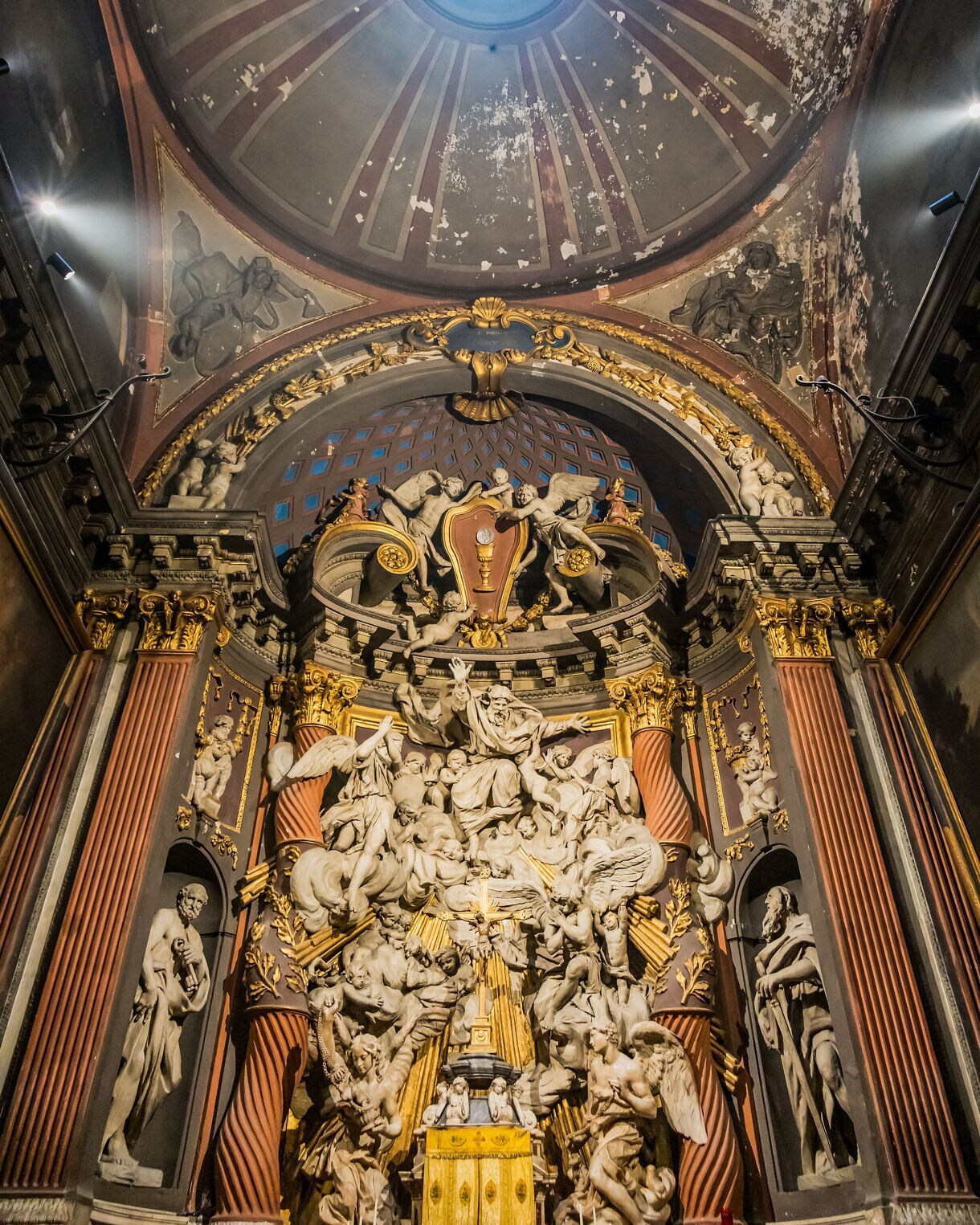 Ornate Baroque altar inside a chapel, featuring a dense sculptural scene of angels and figures in white stone with gold highlights, framed by tall columns and a weathered domed ceiling with a round skylight.