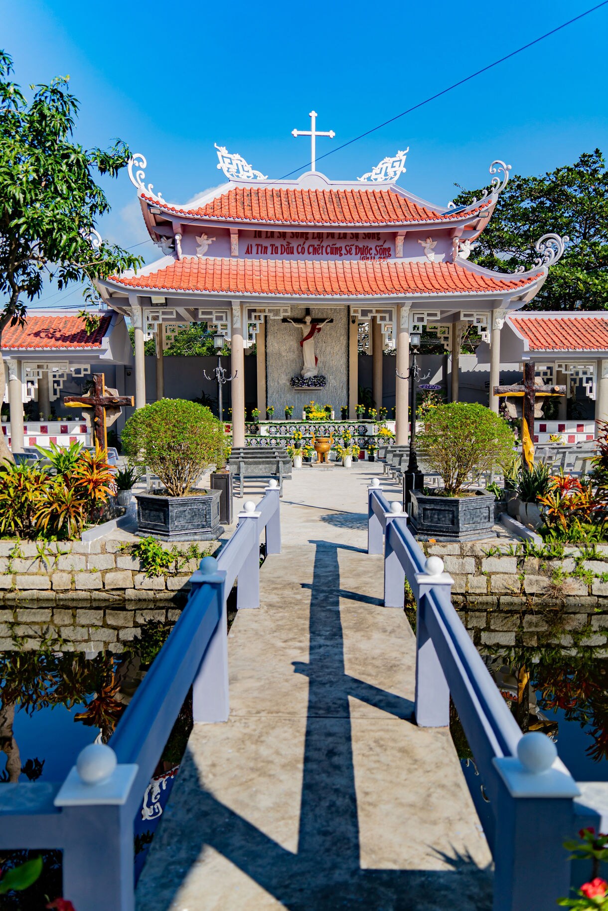 Ornate Catholic cathedral with red-tiled roof and courtyard garden in Nha Trang, Vietnam, viewed from a small bridge over a reflecting pond.