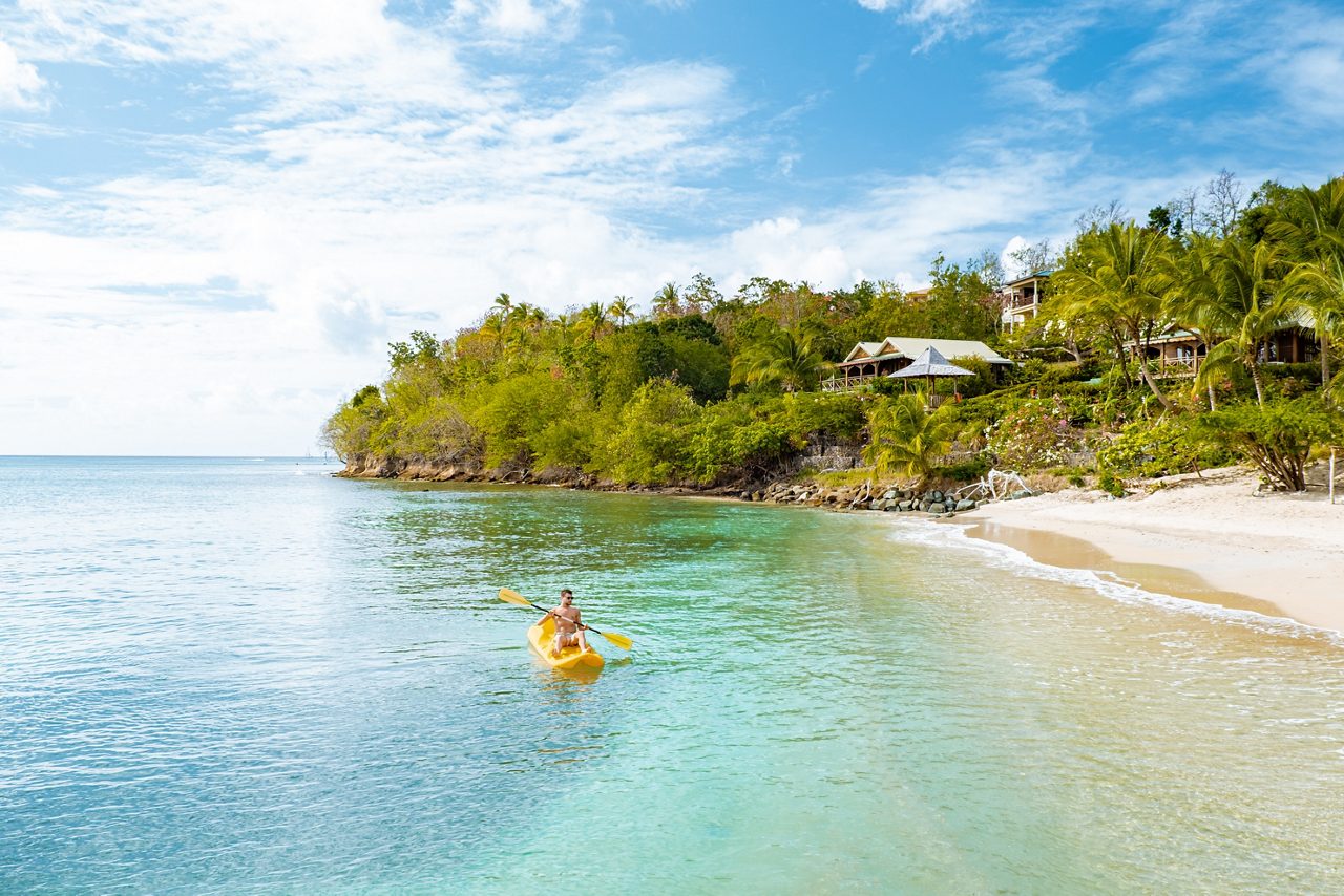 Person kayaking in bright yellow kayak on crystal-clear turquoise water at a tropical beach with white sand, lush green vegetation, and vacation homes nestled among palm trees under a blue sky with white clouds.
