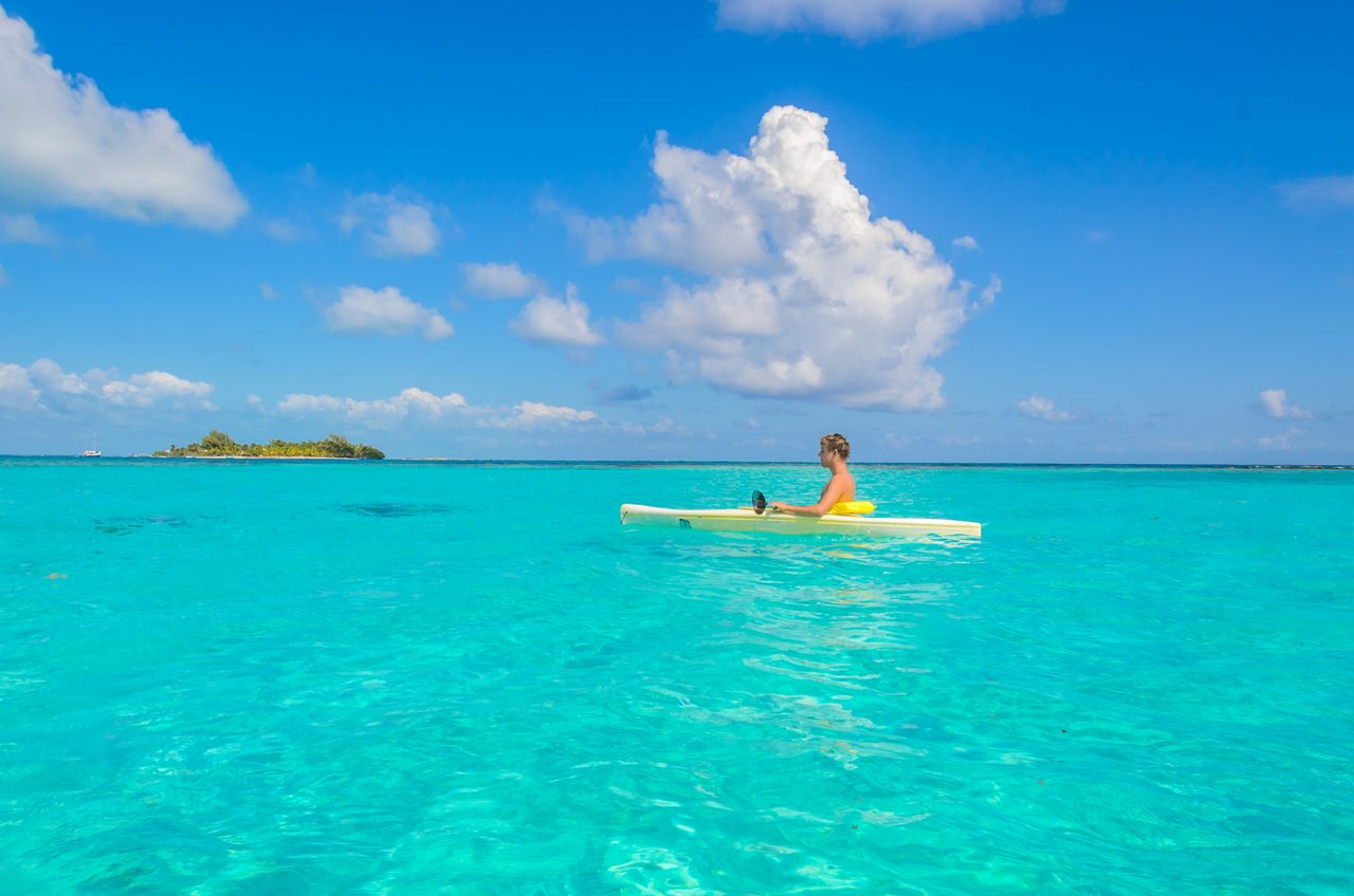 A person kayaking in the turquoise waters of Cays, with a peaceful island in the distance under a bright blue sky.