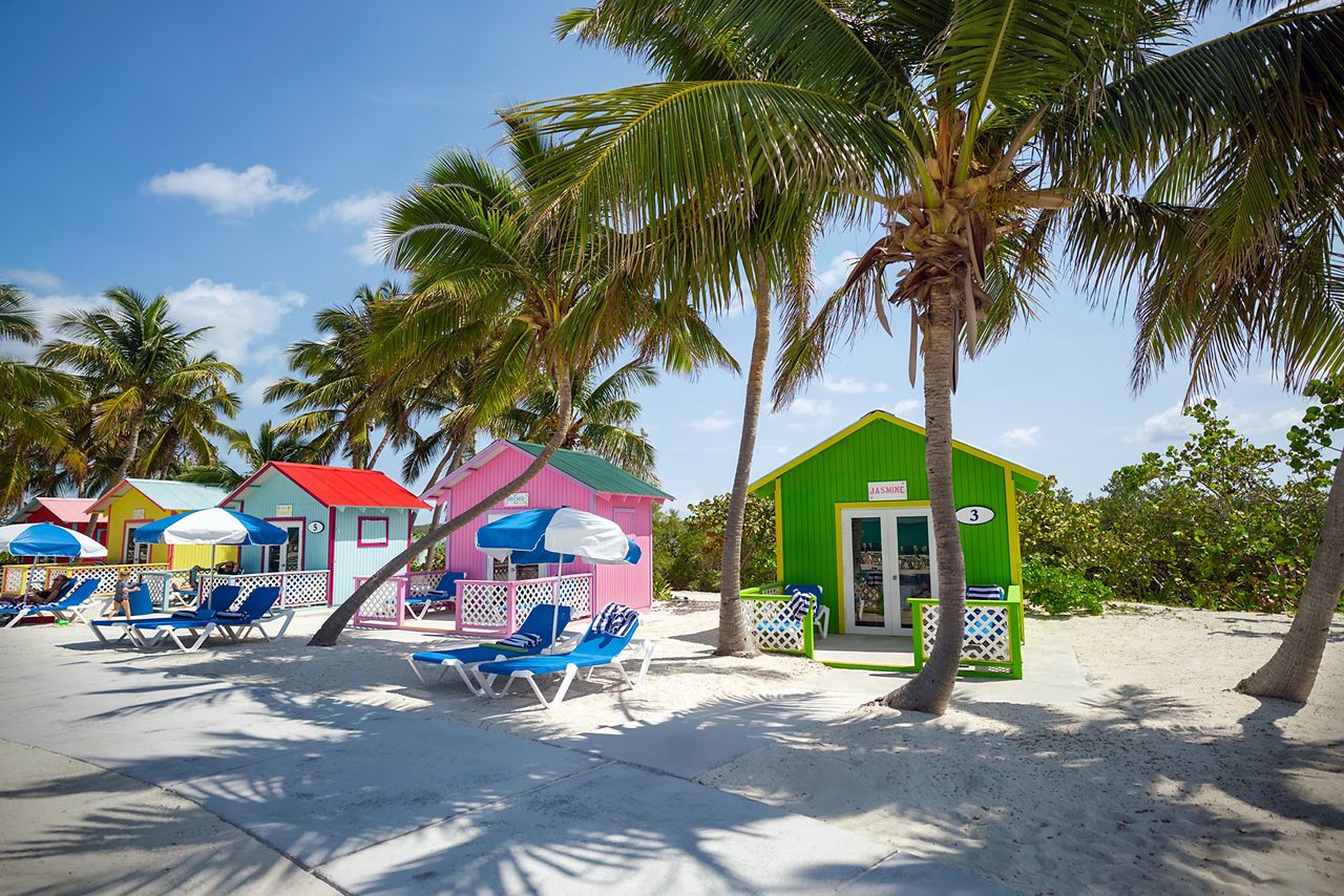 Colorful private beach bungalows with red, pink, green and blue roofs, surrounded by palm trees and lounge chairs on the sandy beach.