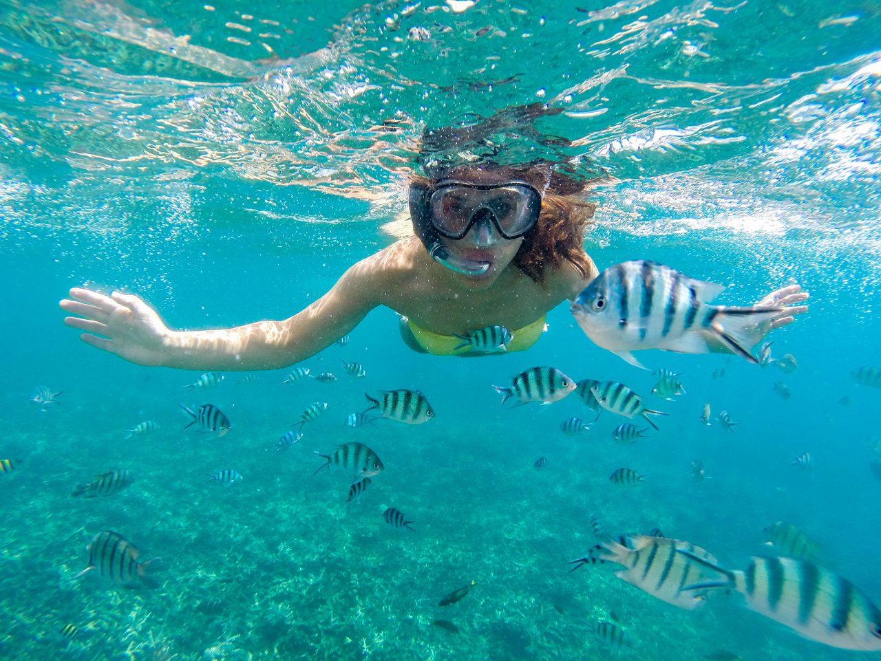 A woman snorkeling underwater, smiling as she swims with striped fish in the clear, turquoise water near Cays.