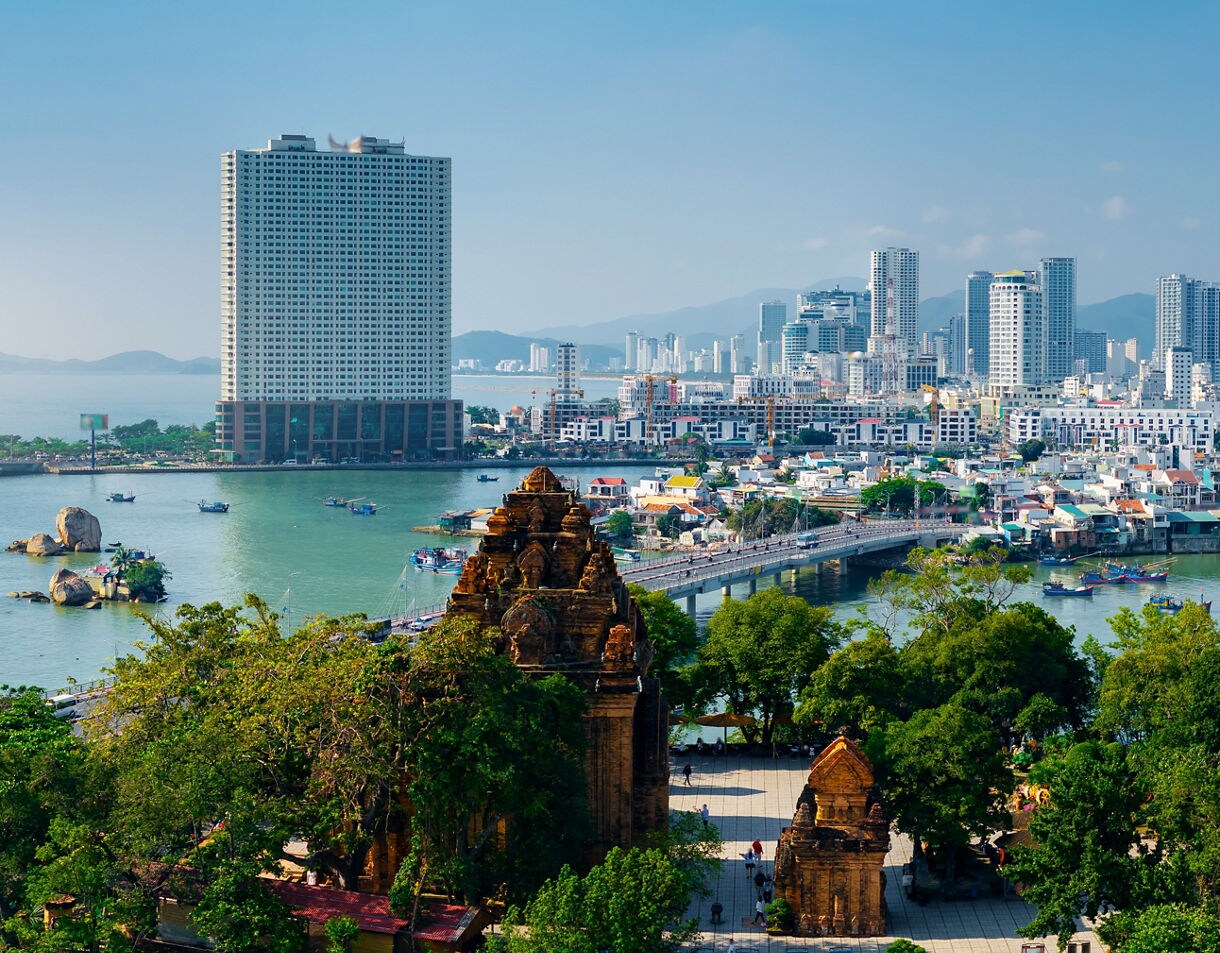 Aerial view of Nha Trang, Vietnam, showing the historic Cham Tower in the foreground and a city skyline of tall buildings along the turquoise bay.