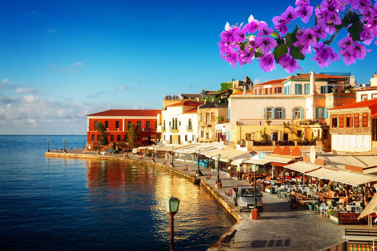 Waterfront view of Chania’s Old Venetian Harbor in Crete, showing colorful seaside buildings, outdoor cafés and the iconic lighthouse under a bright blue sky.