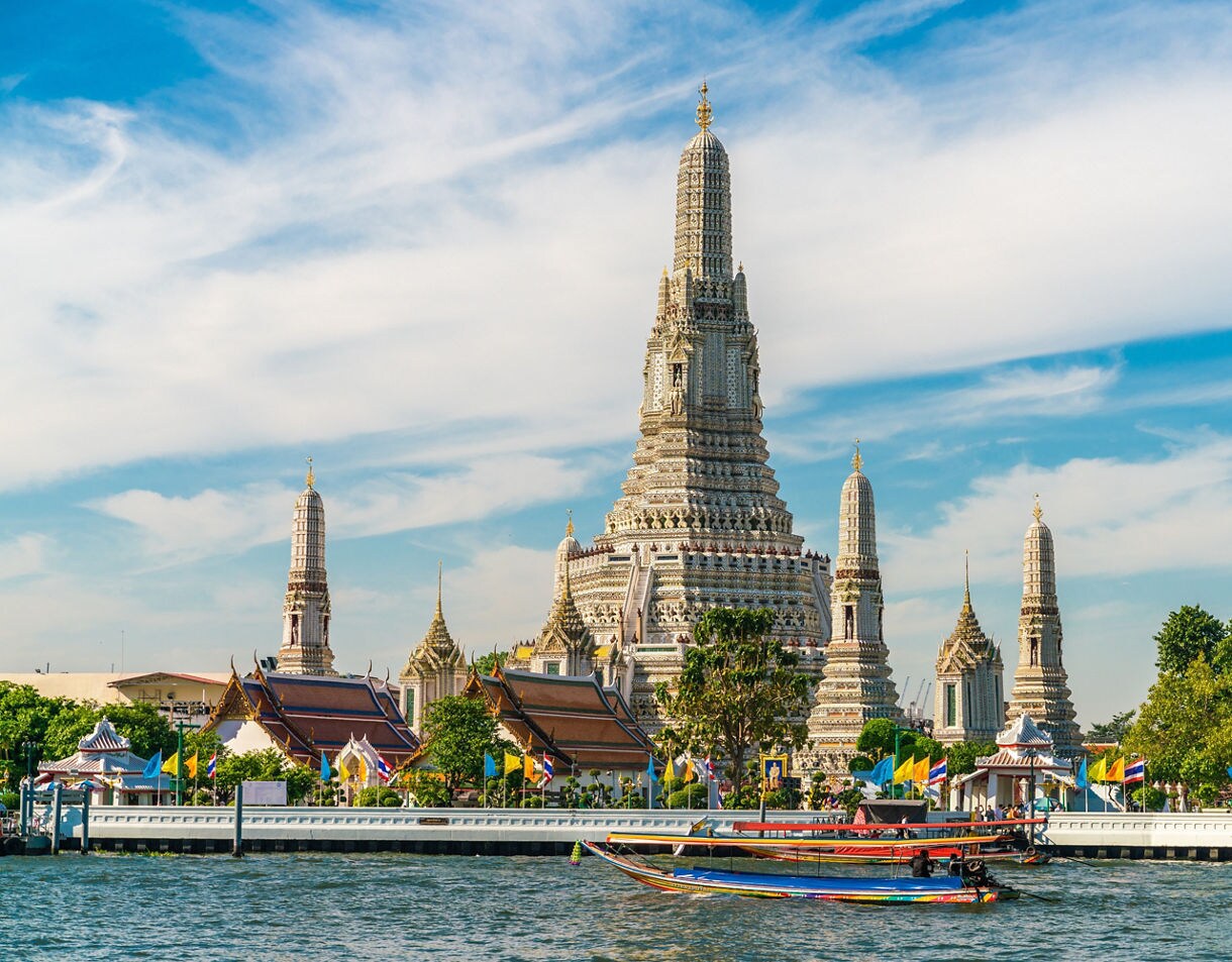 View of Wat Arun temple complex with ornate spires rising behind colorful longtail boats on the Chao Phraya River under a blue sky.