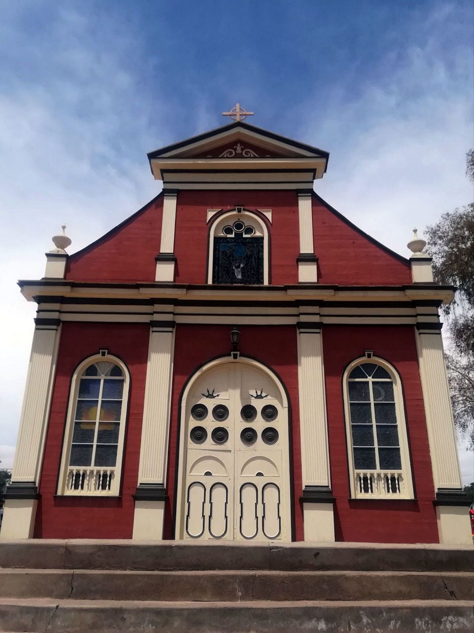 Front view of a small red chapel with cream columns, arched stained-glass windows and a decorative cream door beneath a cross-topped gable.