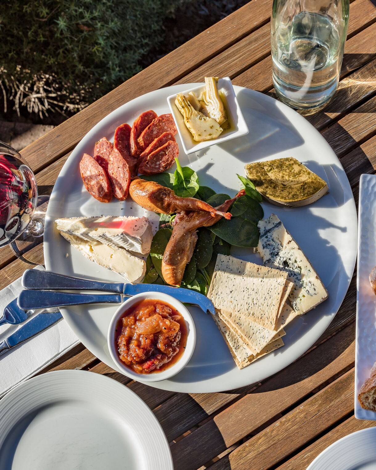 A wooden outdoor table with a charcuterie board featuring assorted cheeses, salami, grilled vegetables, chutney and olives, served with sliced bread, olive oil and a glass of red wine.