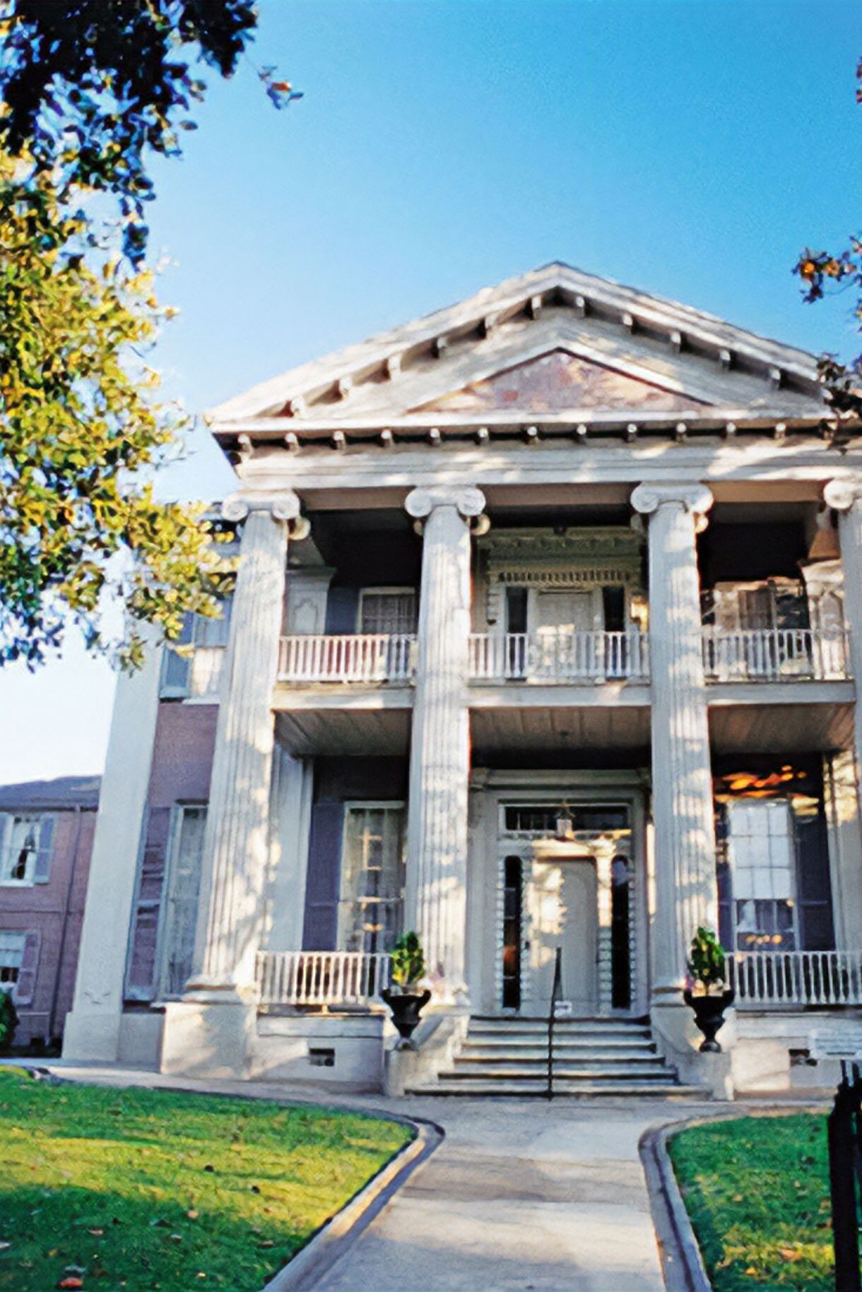 A grand neoclassical building with white columns and a symmetrical façade, surrounded by green trees under a bright blue sky.