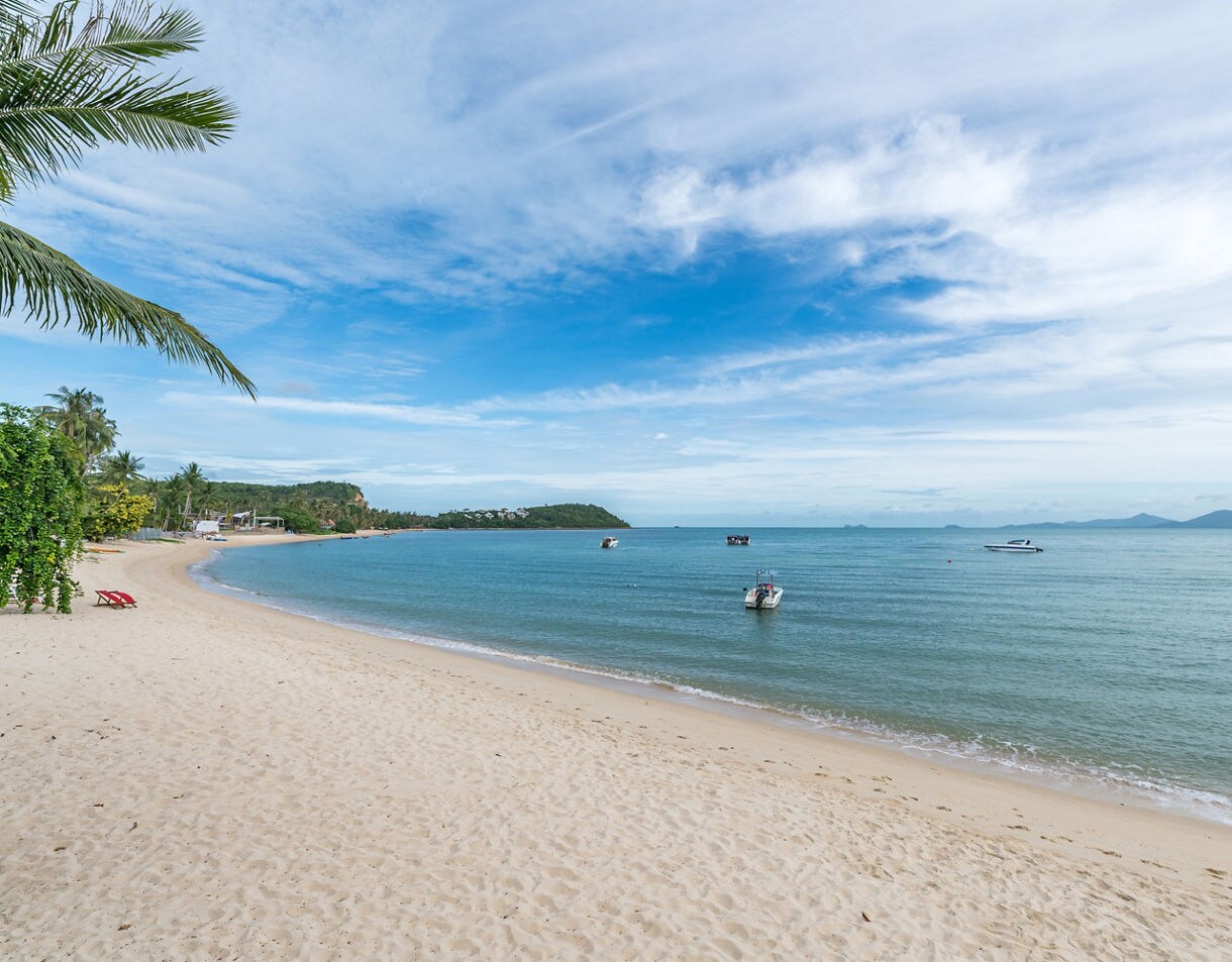 Peaceful view of Chaweng Beach with white sand, gentle waves and small boats floating near the shore on a sunny day.