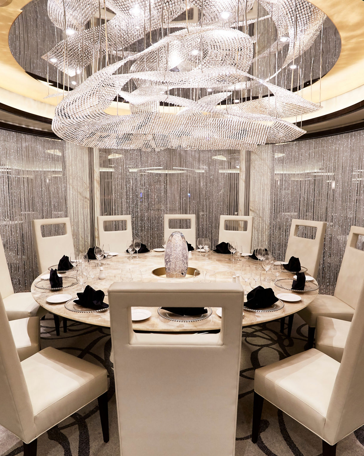 Intimate dining space with crystal chandelier, illuminated curtains and round table featuring sculptural centerpiece where guests enjoy their meal.