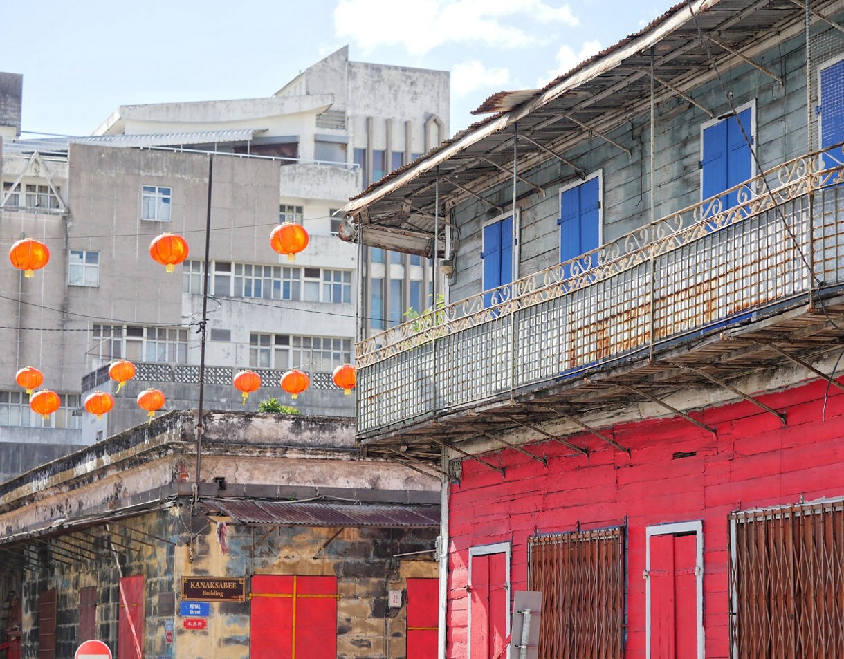 Old buildings with red and blue accents line a street decorated with orange hanging lanterns.