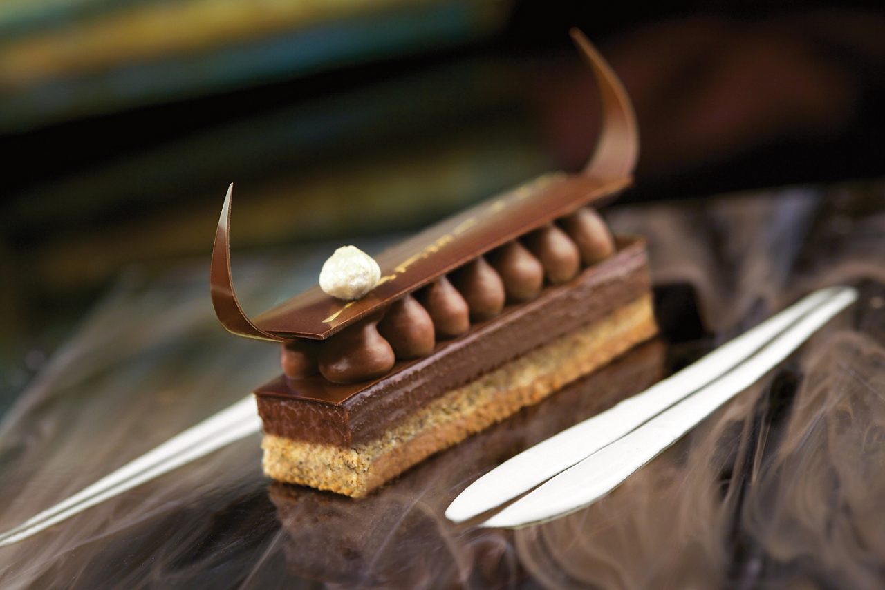 An elegant chocolate hazelnut bar dessert with layers of rich chocolate and a crunchy base, topped with smooth dollops of chocolate ganache, a delicate chocolate garnish, and a touch of citrus cream.