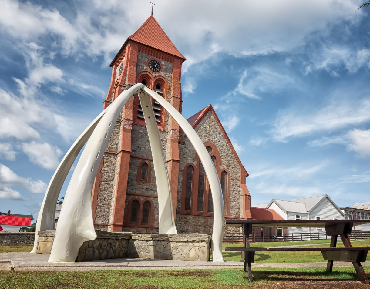 Stone cathedral with red trim behind a large whalebone arch on a grassy lawn under bright blue skies in Stanley.