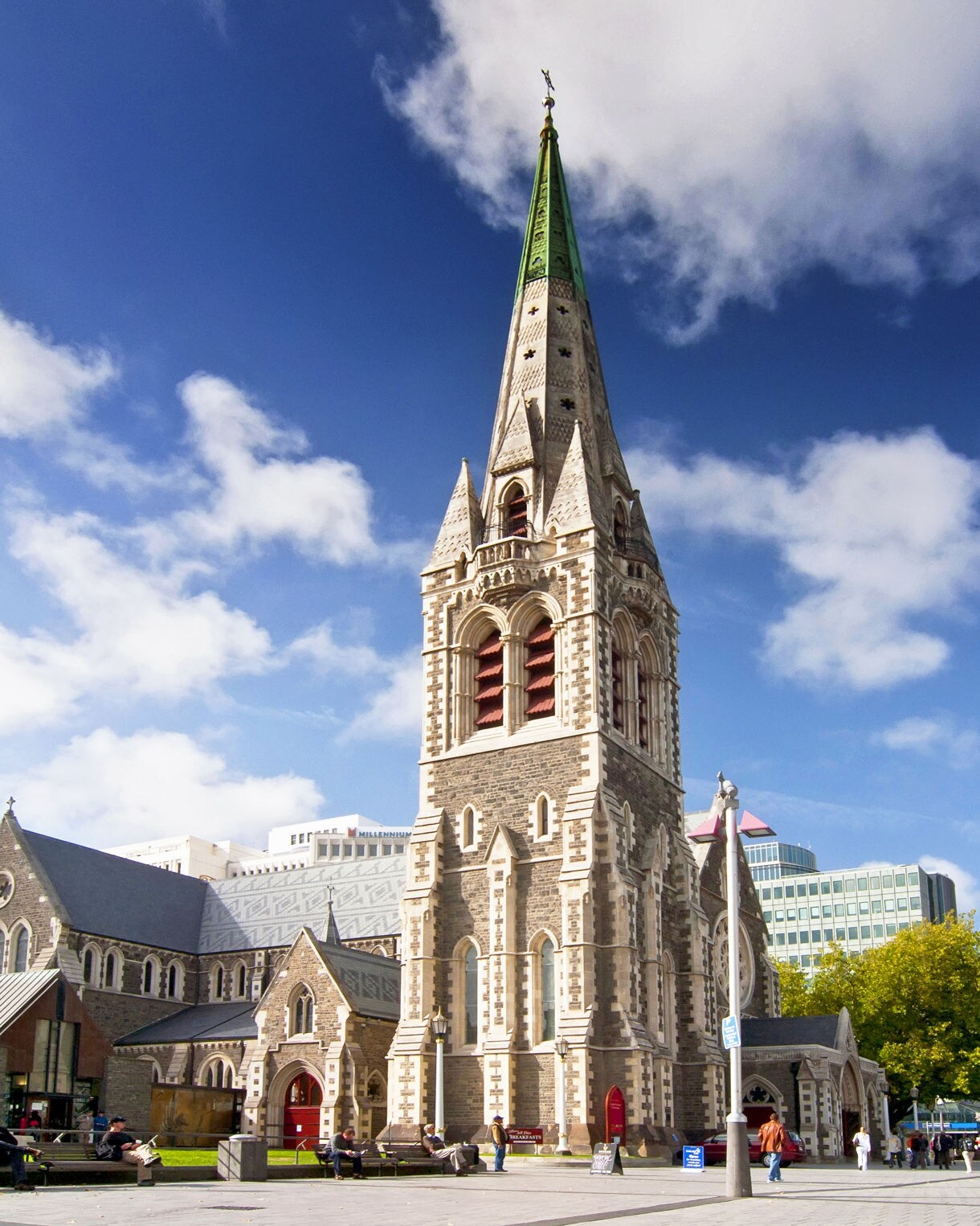 Gothic-style Christchurch Cathedral with its tall stone spire and red doors, surrounded by people walking through Cathedral Square under a bright blue sky.