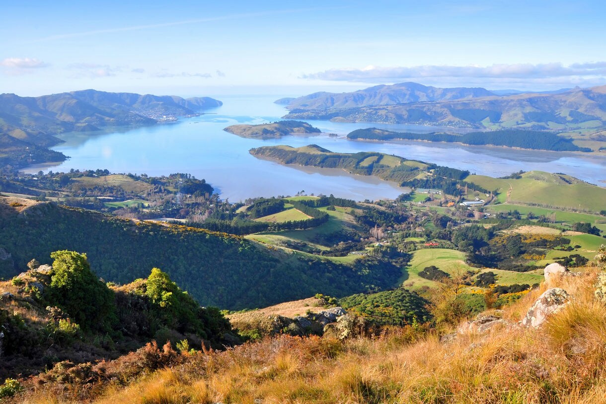 Elevated view of Lyttelton Harbour near Christchurch, New Zealand, showing rolling green hills, calm blue inlets and distant mountain ranges under a clear sky.