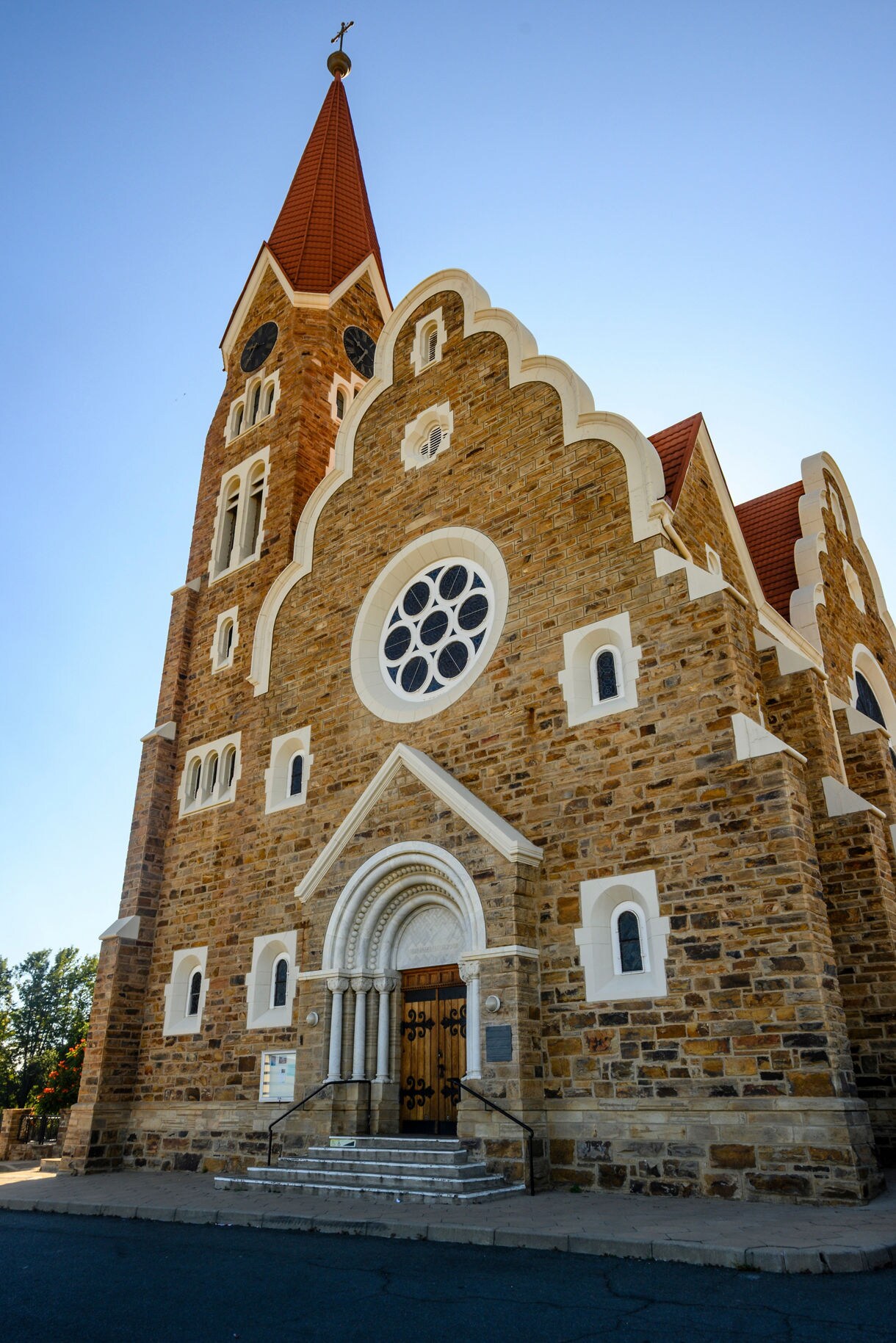 Upward view of Christuskirche in Windhoek, showing its sandstone walls, white trim, arched entrance and tall red-roofed steeple set against a clear blue sky.