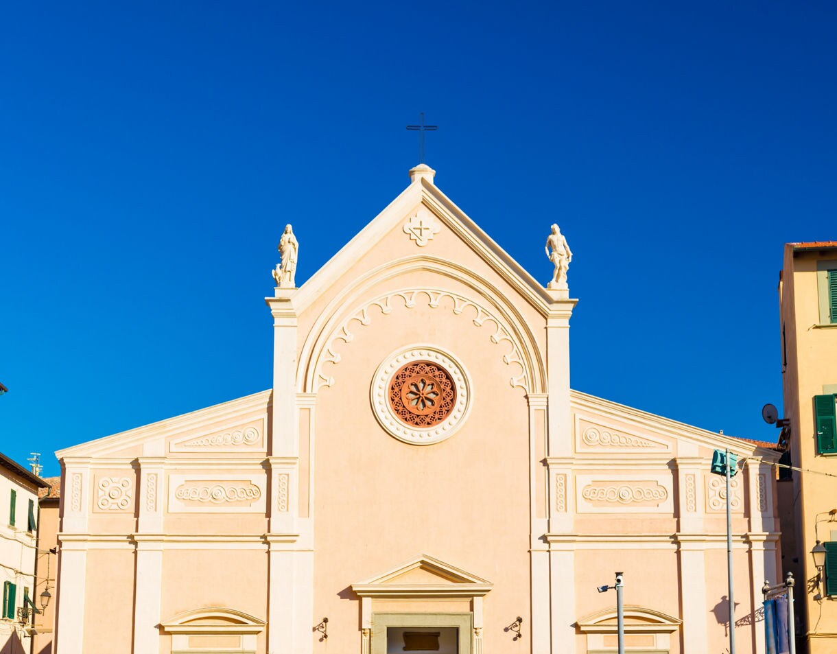 Cream-colored facade of the Church of the Nativity in Capoliveri set against a vivid blue sky with simple statues and green-shuttered homes framing the scene.