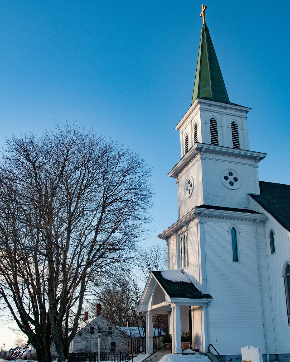 White church with a tall green steeple and gold cross, surrounded by bare winter trees and deep snowbanks along a small town street.