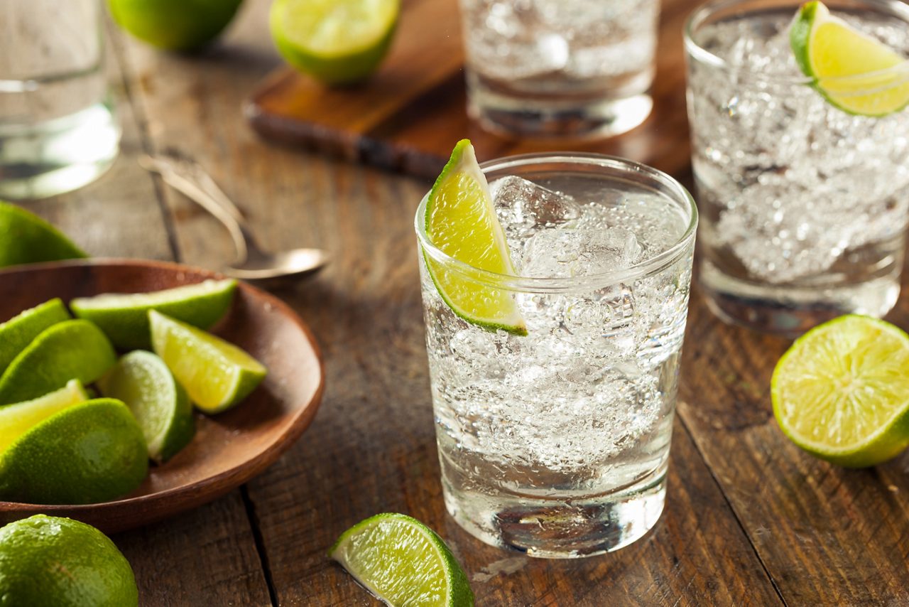 A clear cocktail with lime garnish and ice cubes in highball glass surrounded by fresh limes on wooden table.  