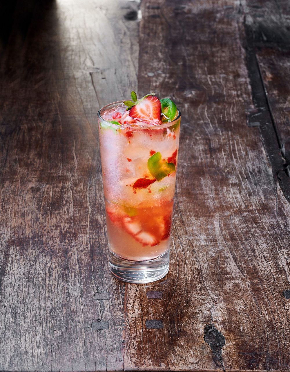 A glass of pink cocktail with strawberry slices, lime wedges and ice on weathered wood surface.