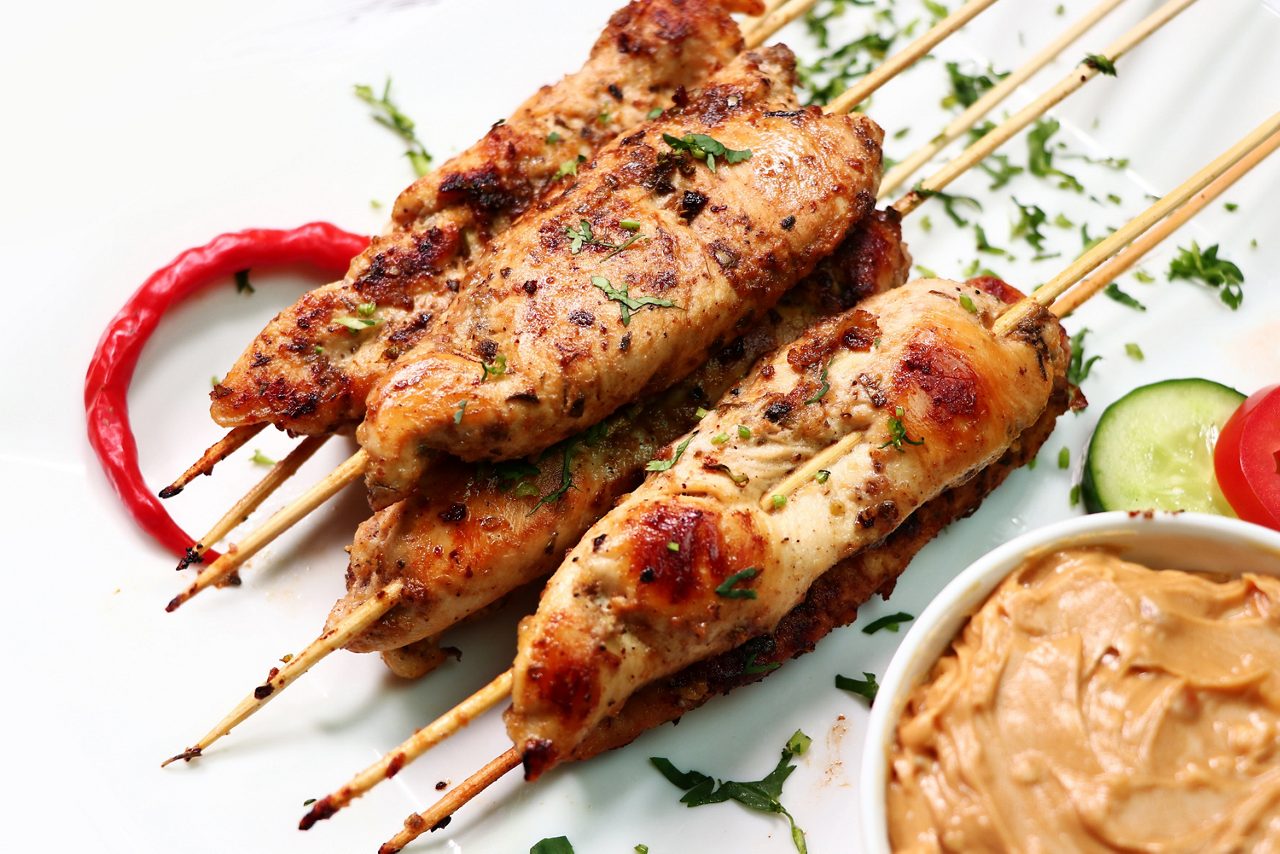 A grilled chicken skewers garnished with herbs, served alongside chili pepper and dipping sauce on white plate.