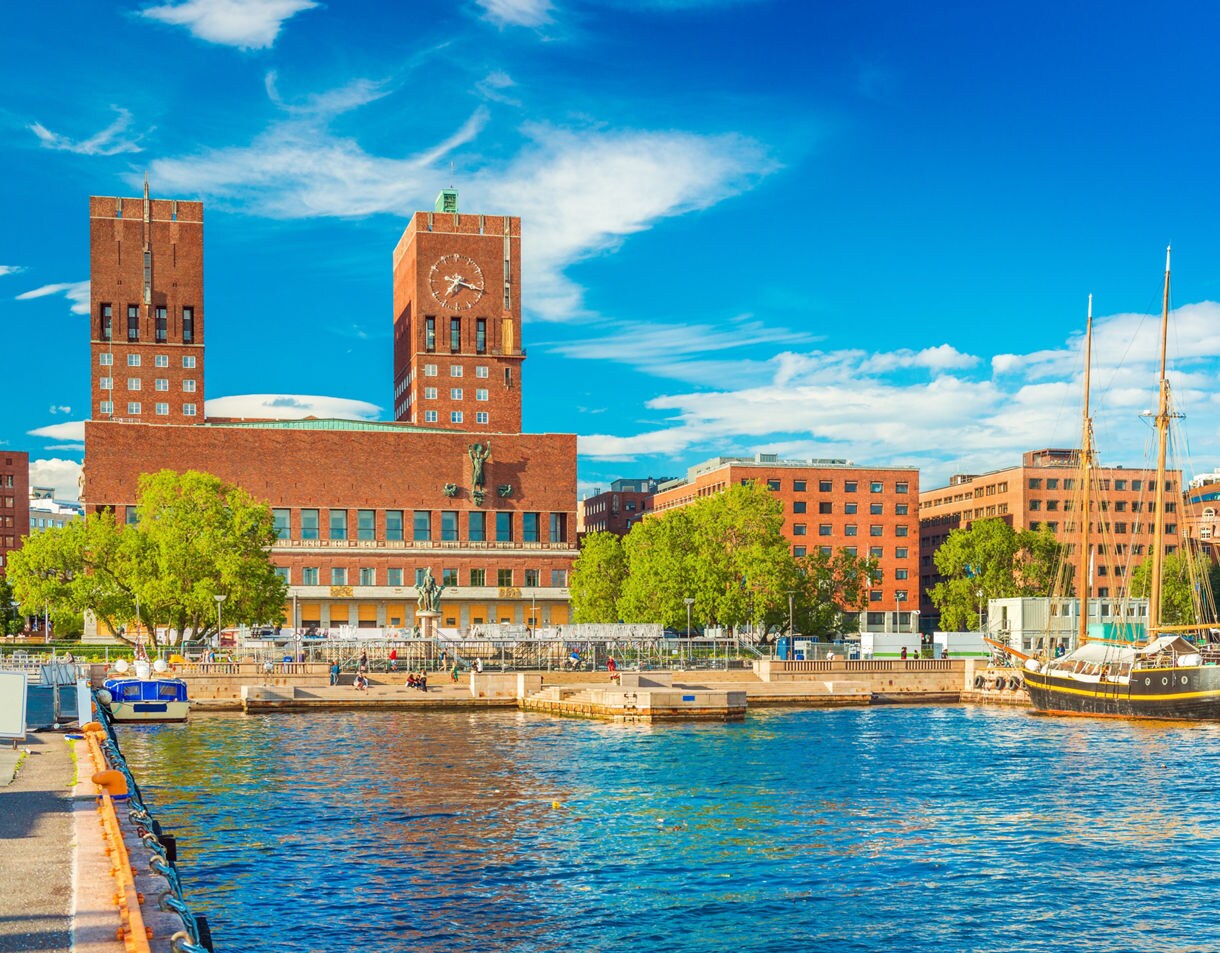 Waterfront view of Oslo City Hall, a red-brick building with twin towers and a large clock, beside boats docked along the harbor on a sunny day.