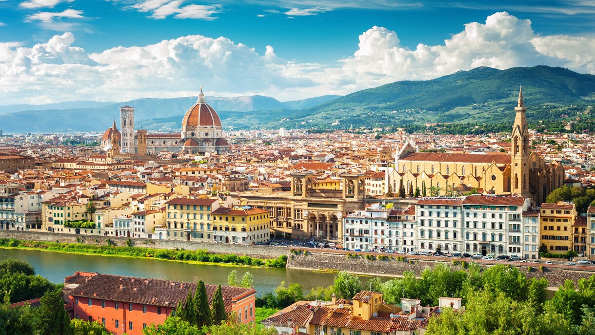 A panoramic view of Florence, Italy, highlighting the Florence Cathedral's red dome, historic buildings, and the Arno River, with lush hills in the background under a bright blue sky.