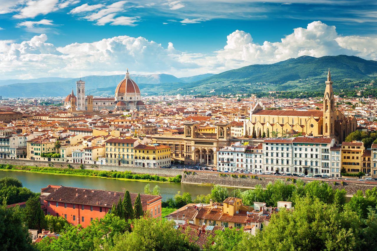 A panoramic view of Florence, Italy, highlighting the Florence Cathedral's red dome, historic buildings, and the Arno River, with lush hills in the background under a bright blue sky.