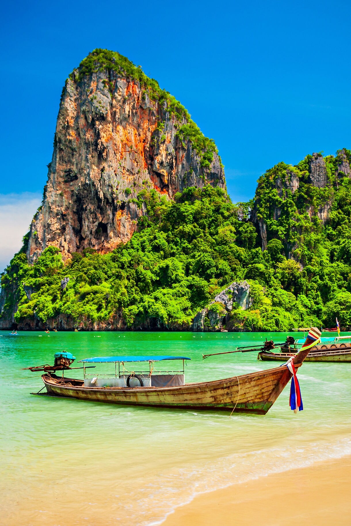 Traditional long-tail boats floating near towering limestone cliffs and clear turquoise waters at a tropical Thai beach.