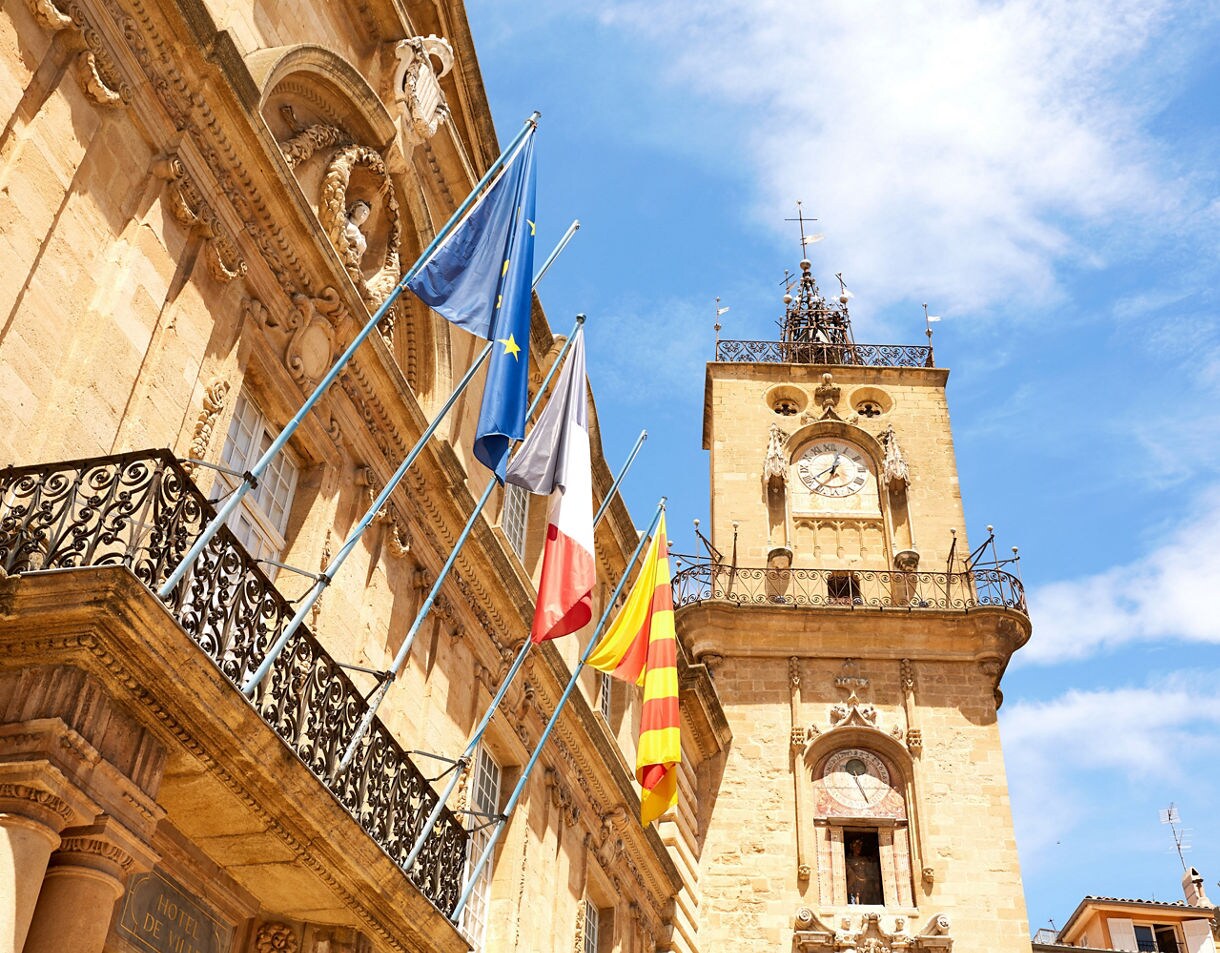 Historic clock tower and town hall in Aix-en-Provence with European and French flags waving against a bright blue sky.