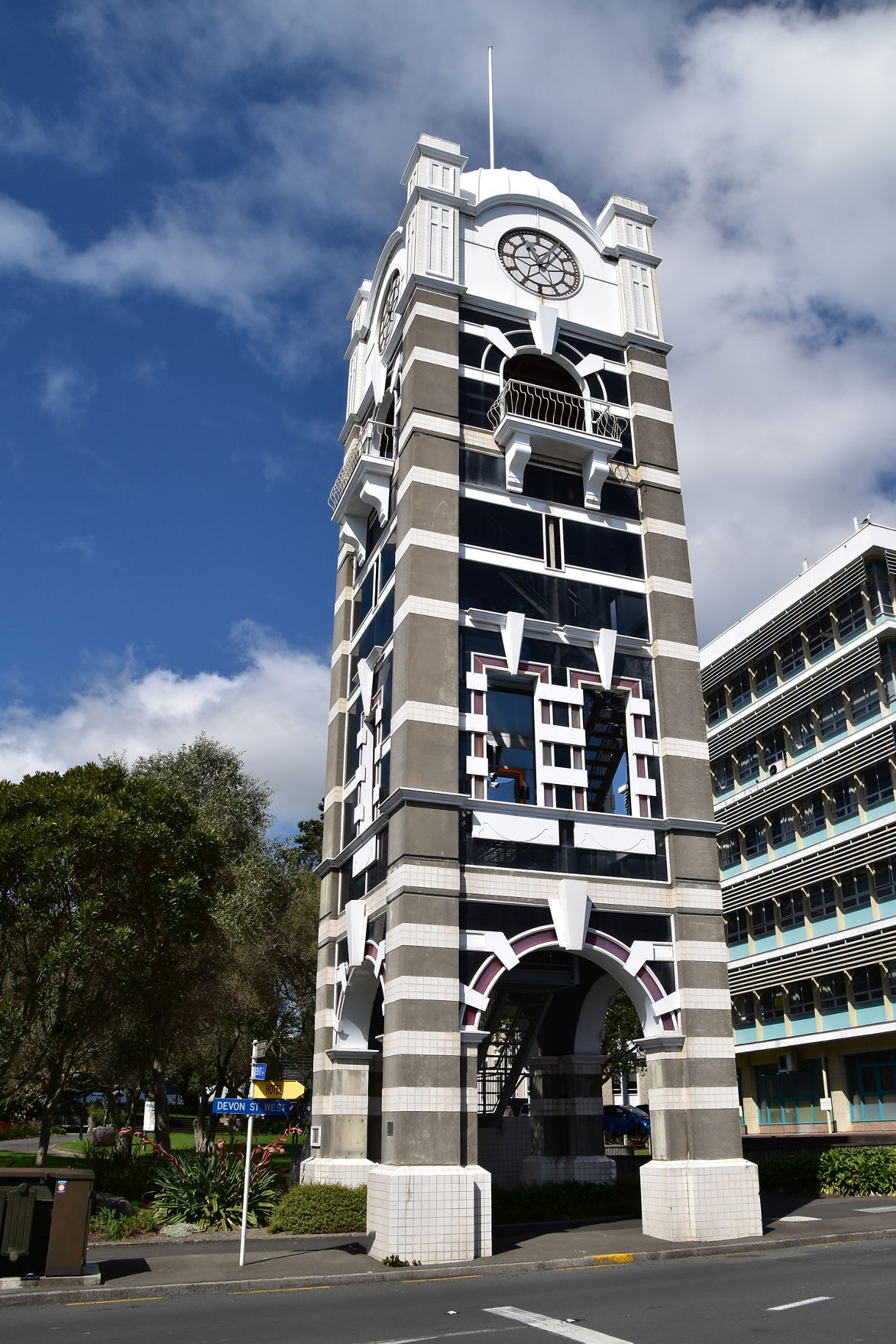 A tall, striped clocktower with arched bases and a decorative clock face, standing along a city street with trees and a modern building nearby under a partly cloudy sky.