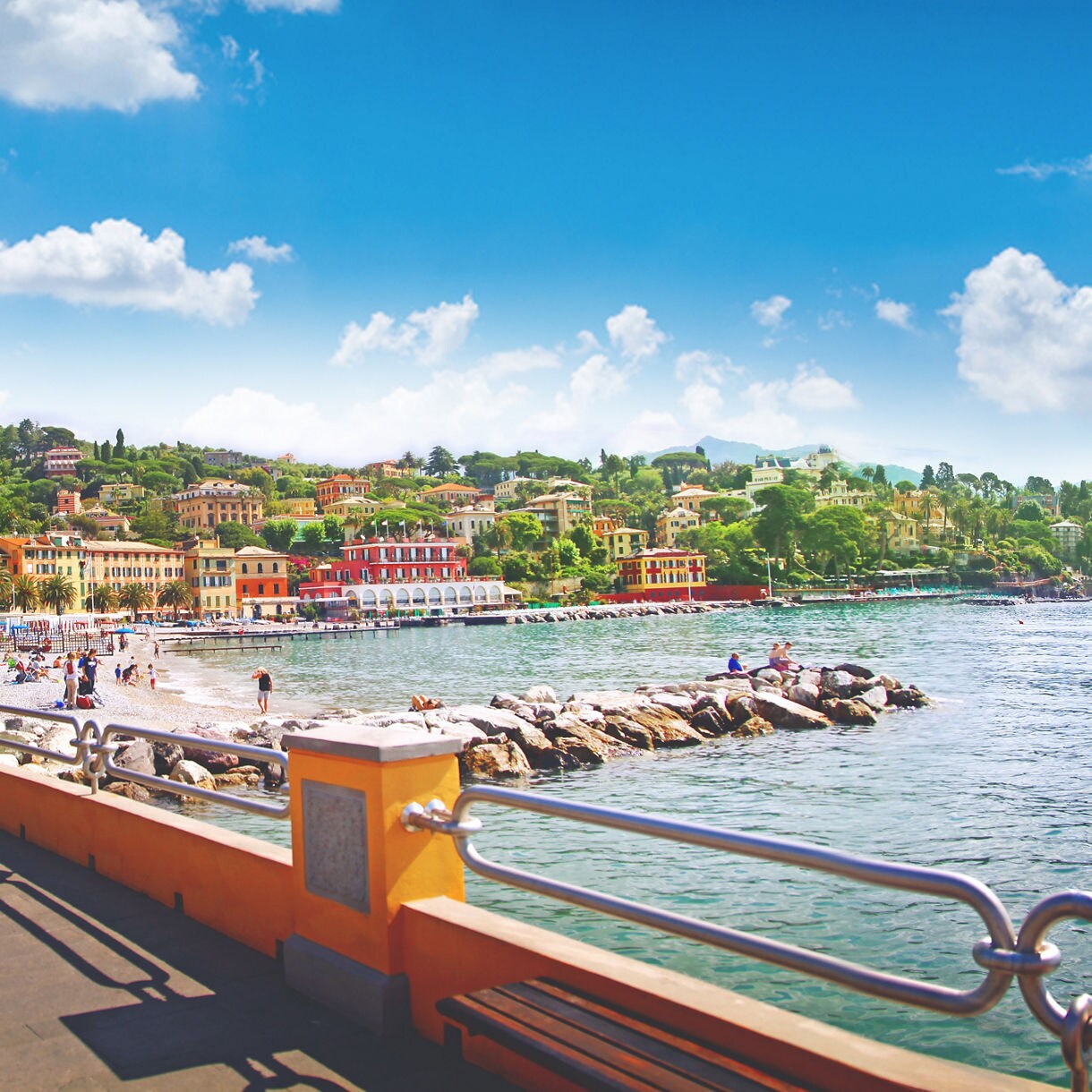 Seaside promenade overlooking a pebble beach and colorful hillside buildings beside calm blue water.