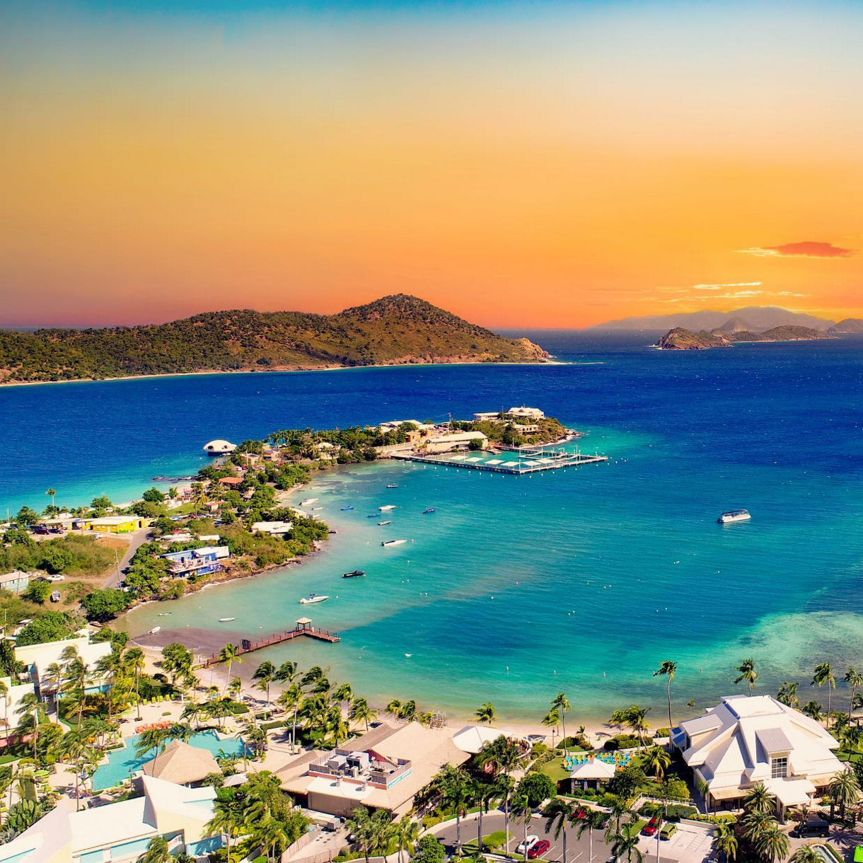 Aerial view of a coastal area at sunset with turquoise waters, scattered buildings, and lush greenery. Hills and islands are visible in the distance under a colorful sky.