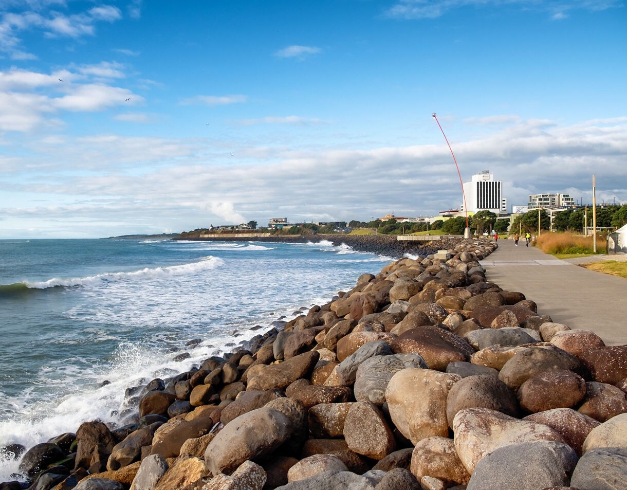 Rocky shoreline along New Plymouth’s Coastal Walkway with waves breaking against large stones and city buildings visible in the distance under a bright blue sky.