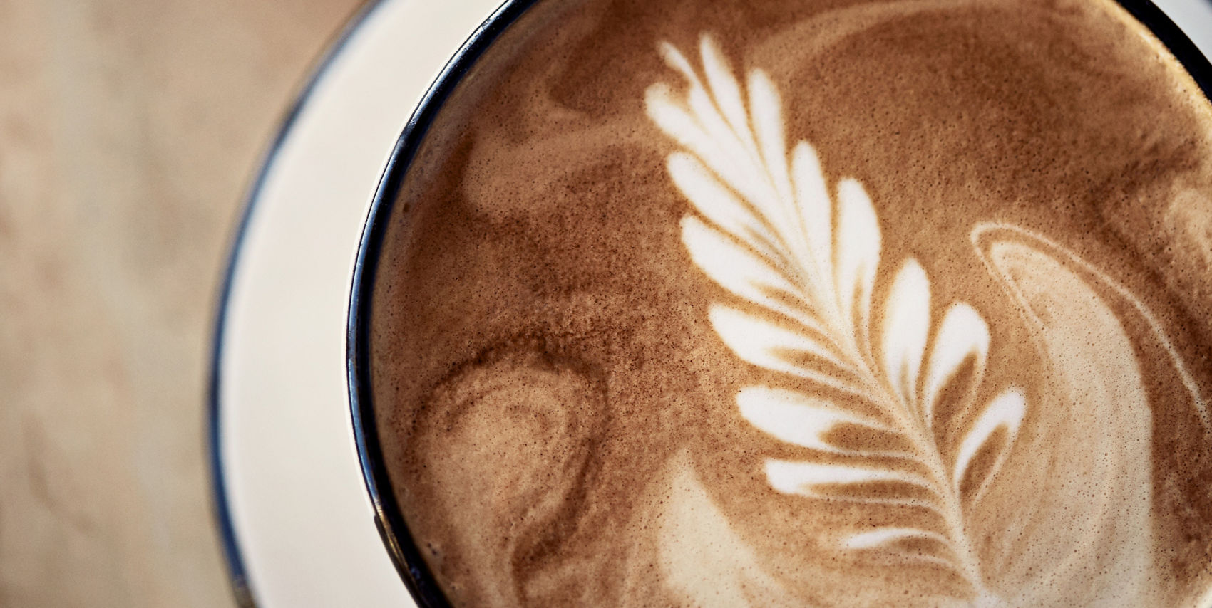 A latte with delicate leaf latte art in a dark ceramic mug, beside a plate with a baked pastry.