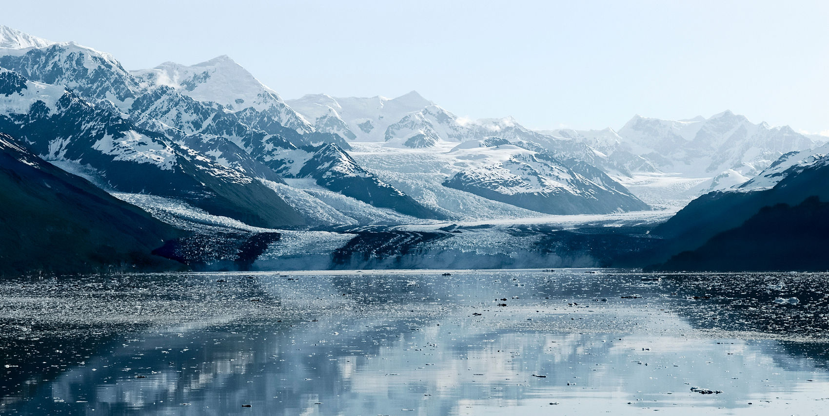 A wide view of College Fjord showing multiple glaciers flowing between snow-covered mountains toward dark blue water dotted with floating ice chunks.