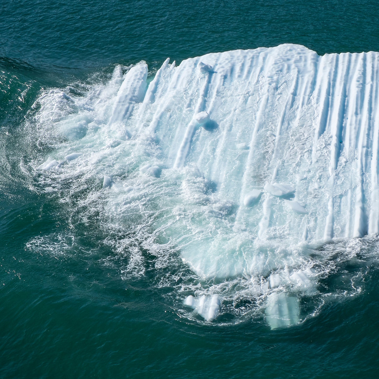 A close-up view of a glacier calving in College Fjord, with large ice chunks breaking off and splashing into the ocean, surrounded by vibrant blue water.