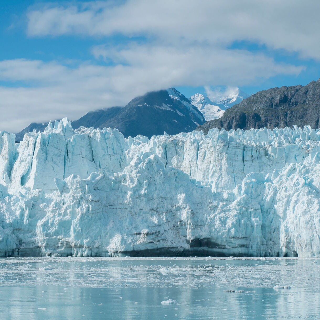 A close-up view of a massive glacier in College Fjord, with jagged ice formations and stunning blue tones set against the backdrop of snow-capped mountains and a clear sky.