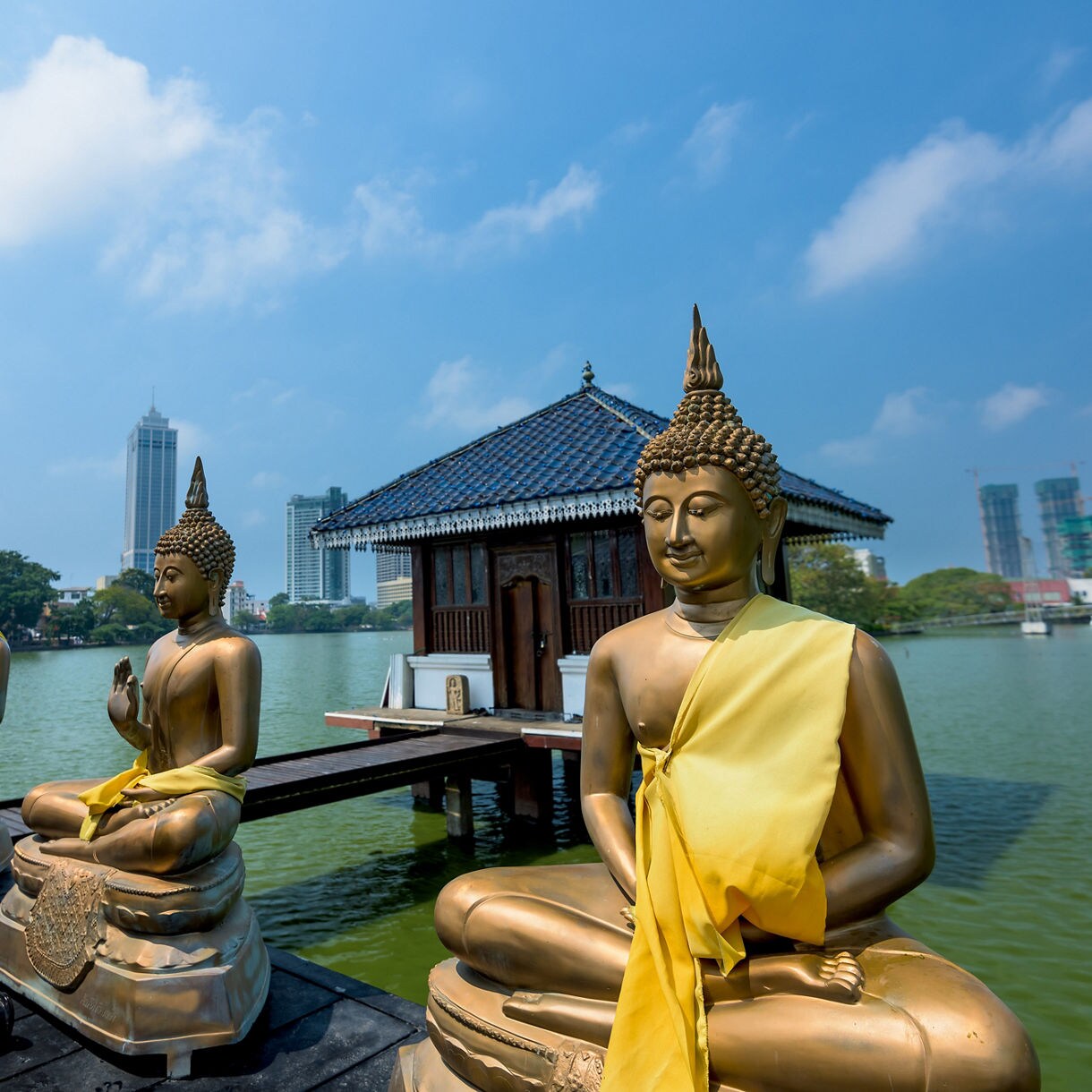 Two golden Buddha statues adorned with yellow sashes are seated by a serene body of water, with a traditional wooden pavilion in the background and modern skyscrapers visible behind the trees under a clear blue sky.