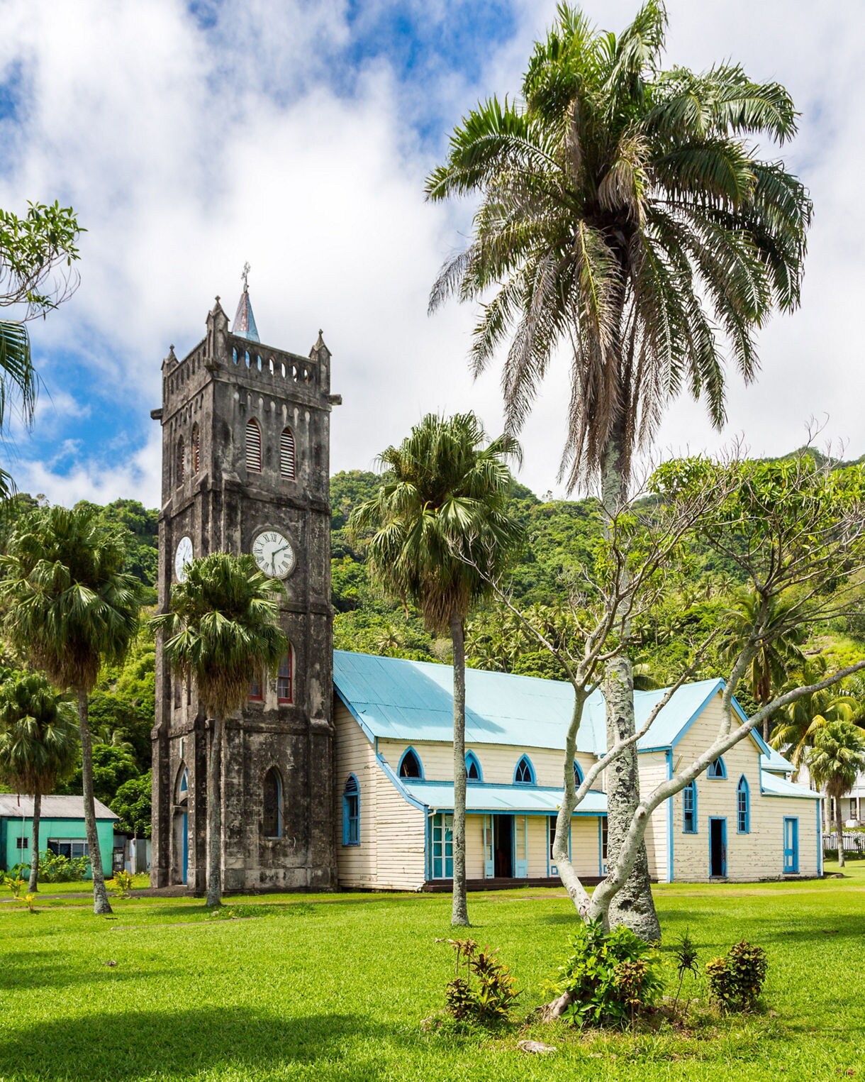 Old stone church with a clock tower and blue roof set on a grassy lawn, surrounded by palm trees and tropical mountains.