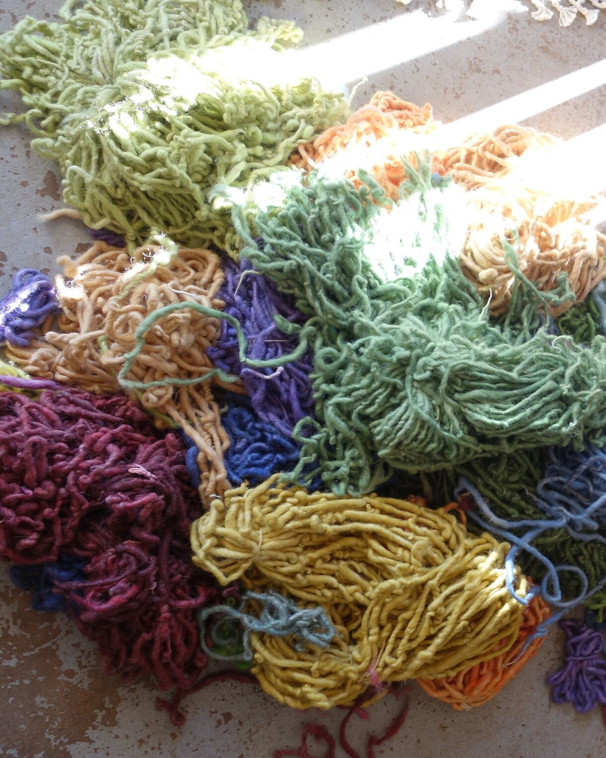 A scattered collection of multicolored wool yarn piles on a sunlit floor, featuring shades of green, yellow, blue, red and purple illuminated by slanted beams of light.