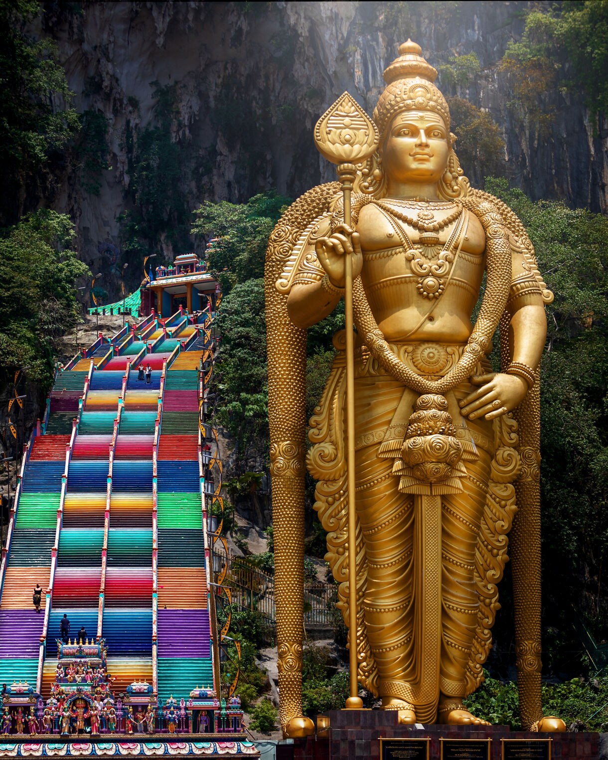 Batu Caves entrance with a giant golden statue of Lord Murugan beside vibrant rainbow-colored steps leading into the limestone cave.