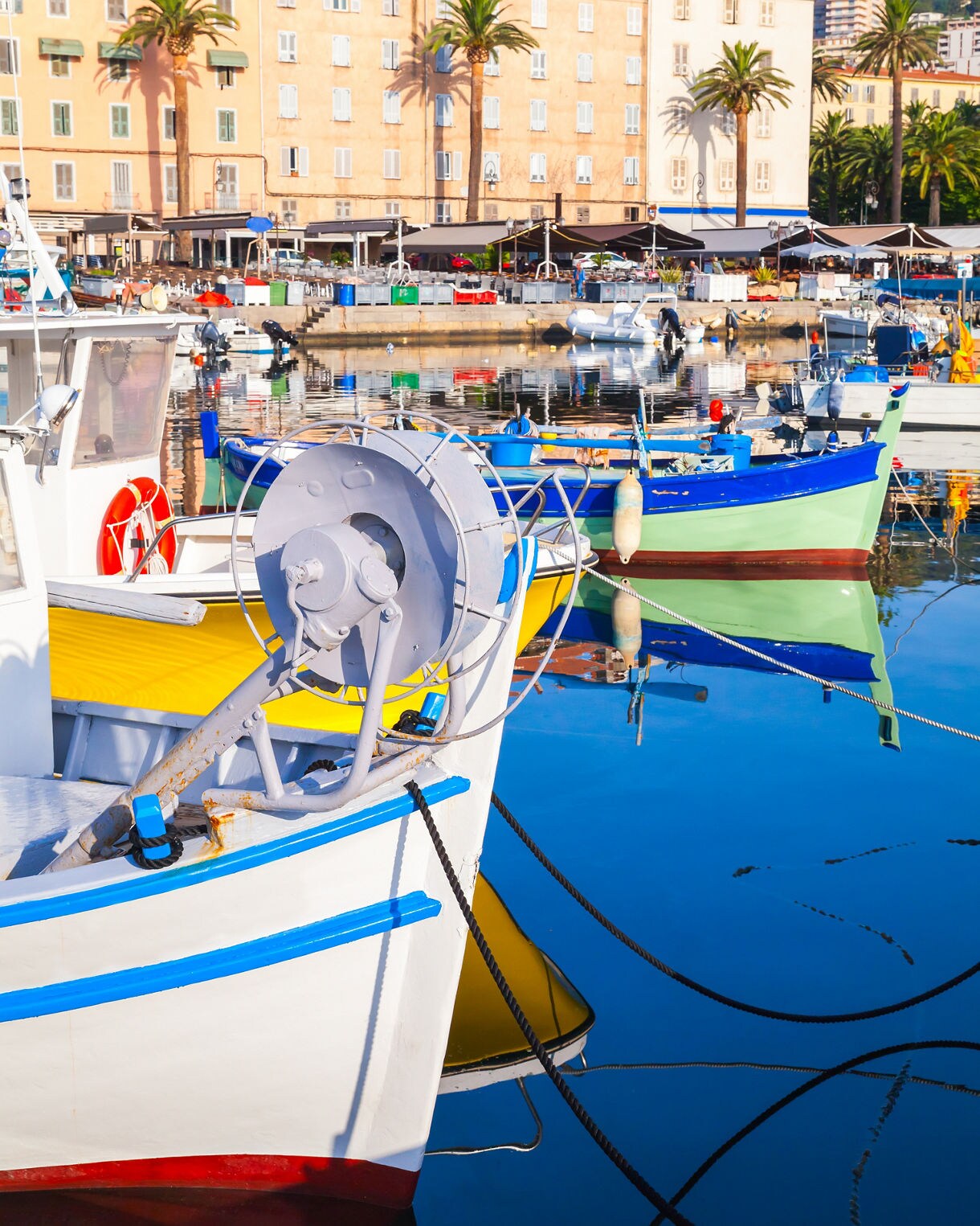 Colorful wooden boats docked in Ajaccio’s harbor, their reflections shimmering on calm blue water beneath palm-lined buildings.