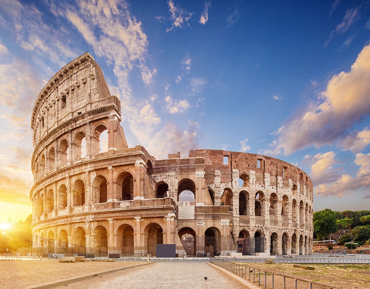 The Colosseum in Rome at sunrise, with warm golden light illuminating its ancient stone arches against a soft blue sky.