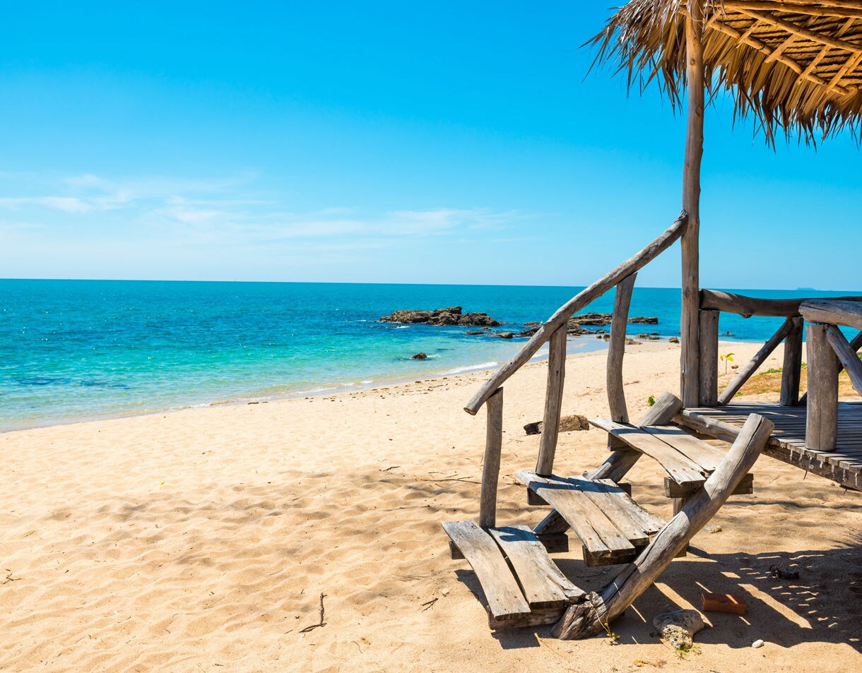 Rustic wooden beach hut with worn steps facing a bright sandy shore and clear blue ocean under a cloudless sky.