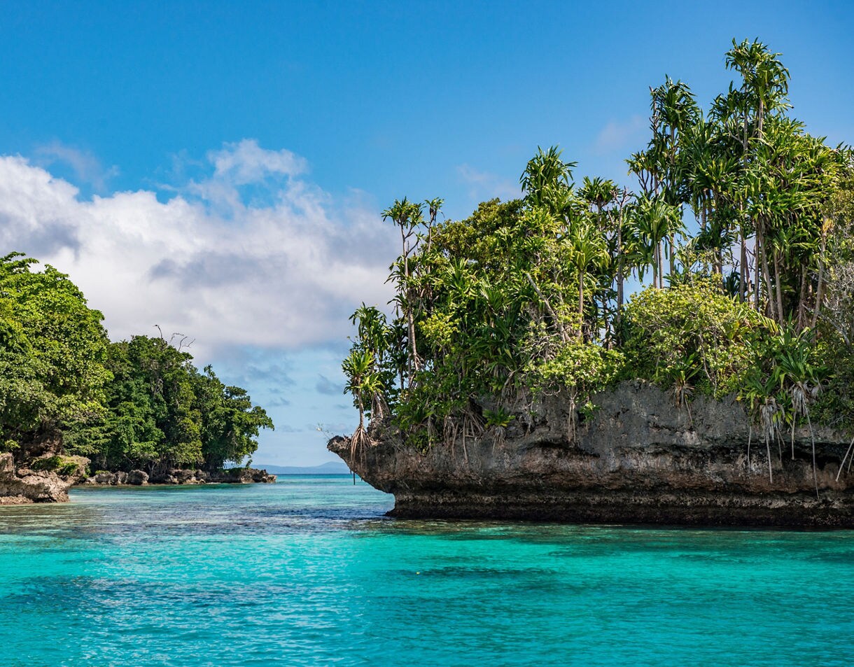 Clear blue water flowing between two rocky, forested islands with bright tropical vegetation under a sunny sky.