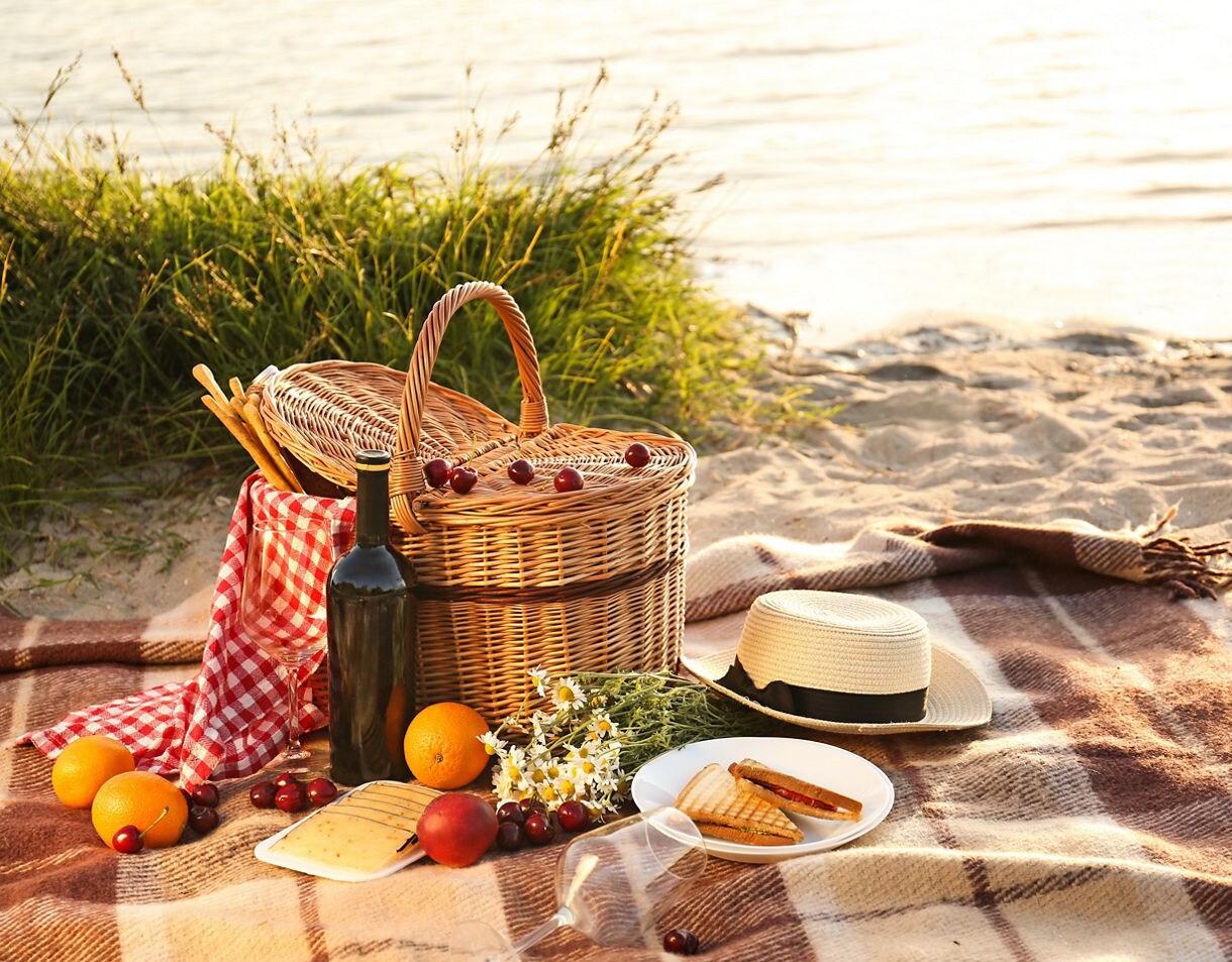 Picnic setup on a plaid blanket with a wicker basket, fruit, cheese, a bottle of wine and a sun hat near sandy shore at sunset.