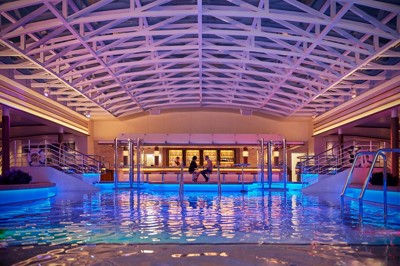 A spacious indoor pool area on a cruise ship, with two people seated at a bar in the background. The scene features a high white-beam ceiling, soft purple and blue lighting, and reflections from the water creating a calm and inviting atmosphere. 